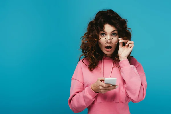 Chica rizada sorprendida tocando gafas y sosteniendo el teléfono inteligente en azul - foto de stock