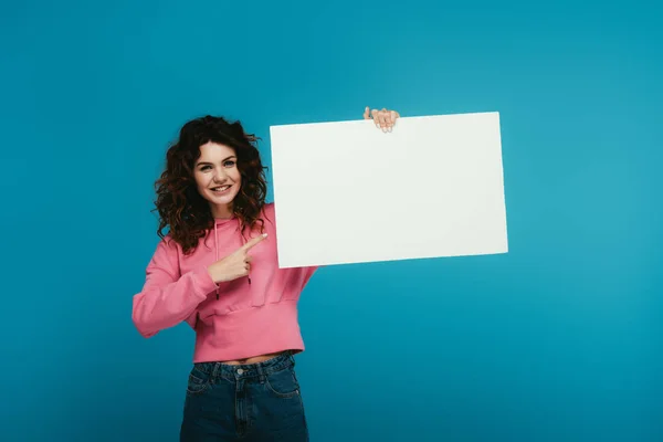 Cheerful curly redhead woman pointing with finger at blank placard on blue — Stock Photo