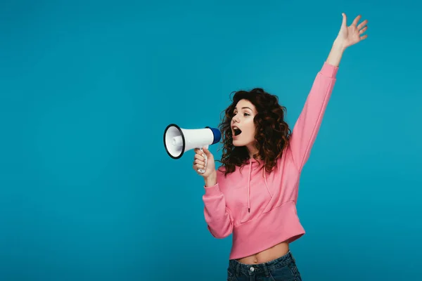 Attractive curly redhead girl screaming in megaphone on blue — Stock Photo