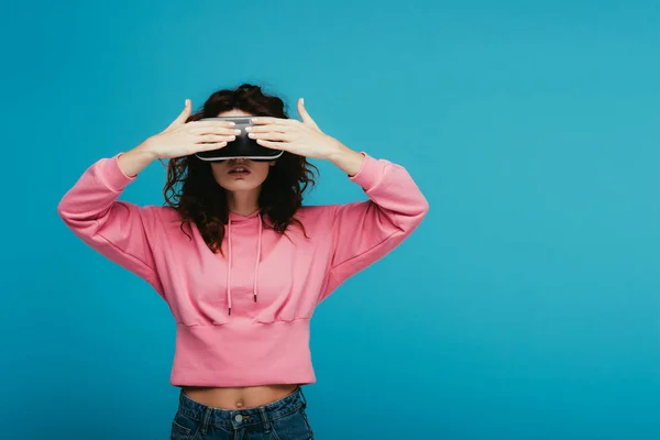 Chica rizada con auriculares de realidad virtual mientras cubre los ojos en azul - foto de stock