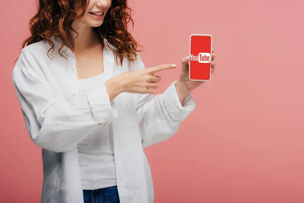 Vista cortada de menina alegre apontando com o dedo para o smartphone com a tela do aplicativo do youtube em rosa — Fotografia de Stock