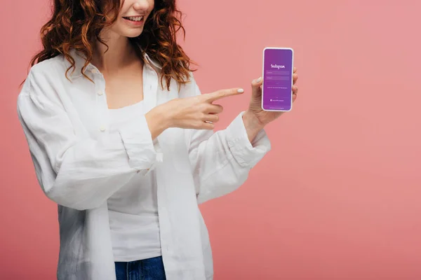 Vista recortada de chica alegre señalando con el dedo en el teléfono inteligente con instagram pantalla de la aplicación en rosa - foto de stock