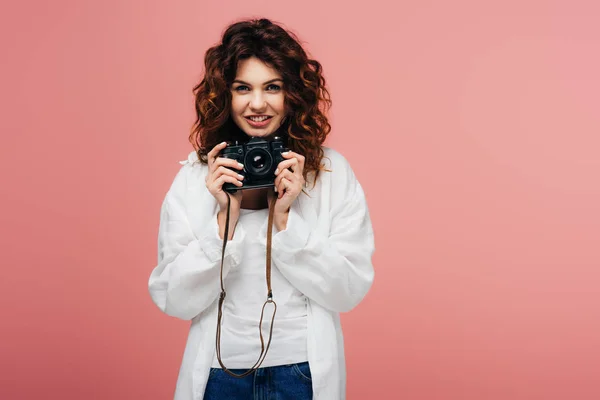 Веселая молодая кудрявая женщина держит цифровую камеру и улыбается на розовый — стоковое фото