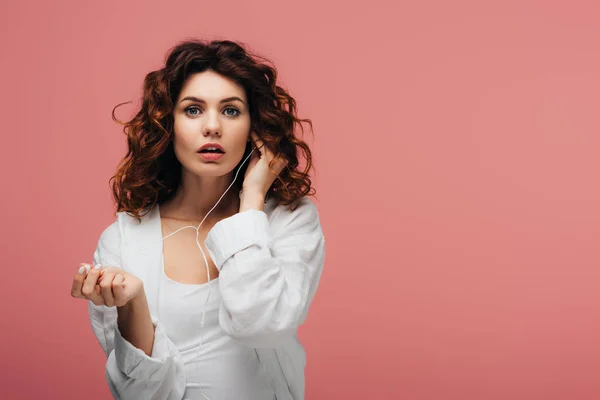 Attractive curly girl listening music in earphones and looking at camera on pink — Stock Photo