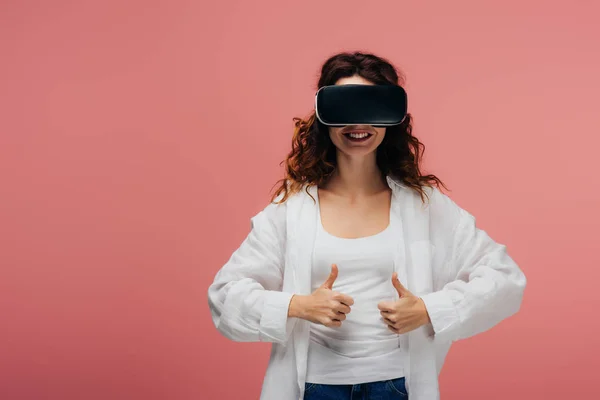 Happy curly redhead woman wearing virtual reality headset and showing thumbs up on pink — Stock Photo