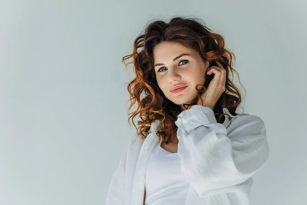 Happy curly young woman touching hair and looking at camera on grey — Stock Photo