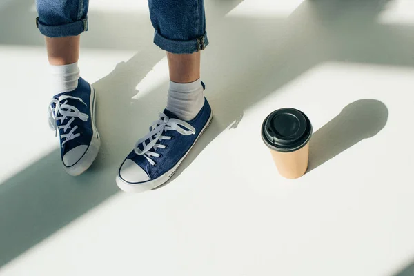 Cropped view of woman in sneakers near paper cup on white — Stock Photo