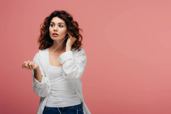 Attractive curly girl with red hair listening music in earphones on pink — Stock Photo