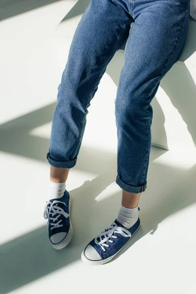 Cropped view of woman in sneakers sitting on white with sunshine — Stock Photo