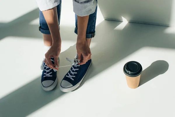 Cropped view of woman tying shoelaces on sneakers while sitting near paper cup on white with sunshine — Stock Photo
