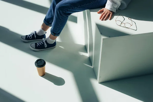 Ausgeschnittene Ansicht einer Frau, die in Turnschuhen neben Pappbecher und Brille auf Weiß mit Sonnenschein sitzt — Stockfoto