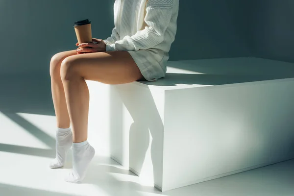 Обрізаний вид молодої жінки, сидячи в шкарпетках і тримаючи паперову чашку на сірому — стокове фото