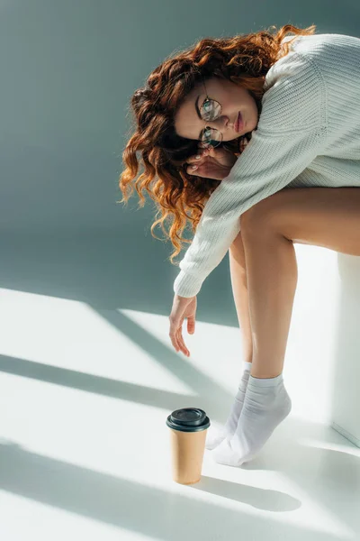 Attrayant fille bouclée dans des lunettes regardant la caméra tout en étant assis près de tasse en papier sur gris — Photo de stock