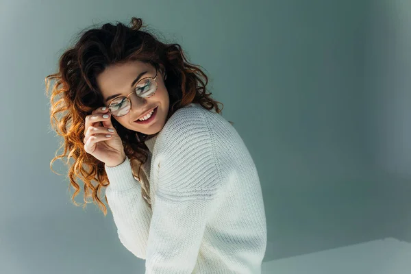 Cheerful curly redhead girl touching glasses and smiling on grey — Stock Photo