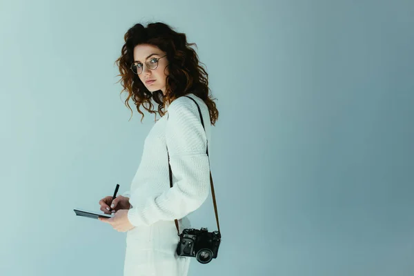 Attraktive junge Frau mit roten Haaren mit Notizbuch und Stift im Stehen mit Digitalkamera auf grau — Stockfoto