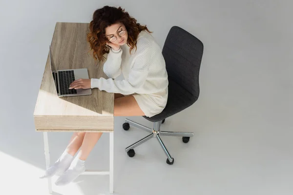 Overhead-Ansicht des lockigen rothaarigen Mädchens sitzt in der Nähe von Laptop auf grau — Stockfoto