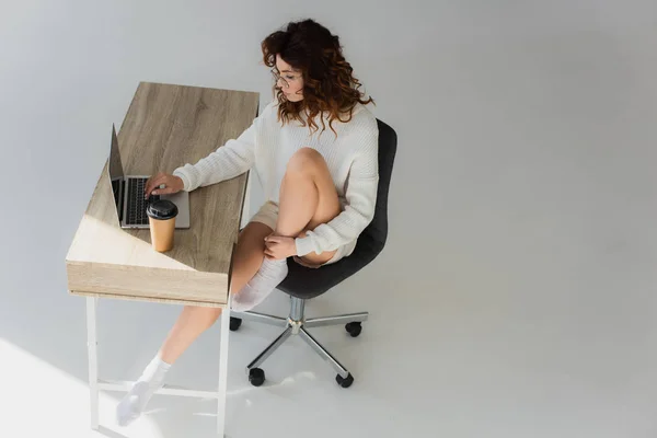 Vista aérea de la mujer rizada en gafas sentado y el uso de la computadora portátil cerca de la taza de papel en gris - foto de stock