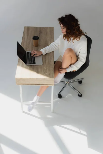 Vista aérea de la mujer joven en gafas sentado y usando el ordenador portátil cerca de la taza de papel en gris - foto de stock