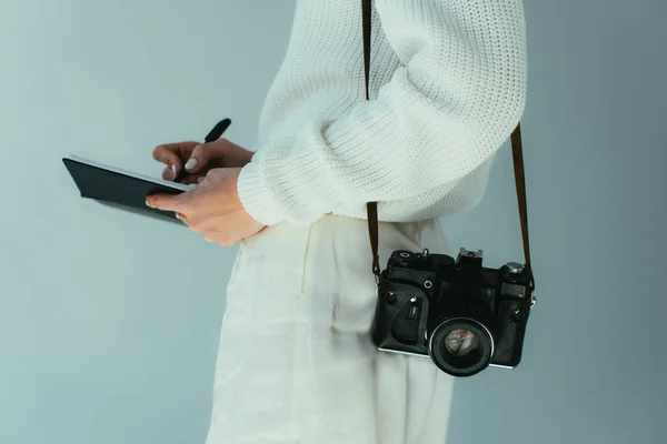 Vue recadrée de jeune femme écrivant dans un carnet tout en se tenant debout avec un appareil photo numérique sur gris — Photo de stock