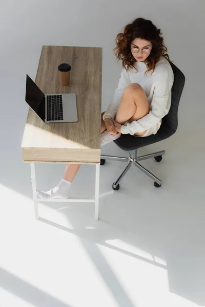 Vista aérea de mujer joven en gafas sentadas cerca de la mesa con portátil y taza de papel en gris - foto de stock