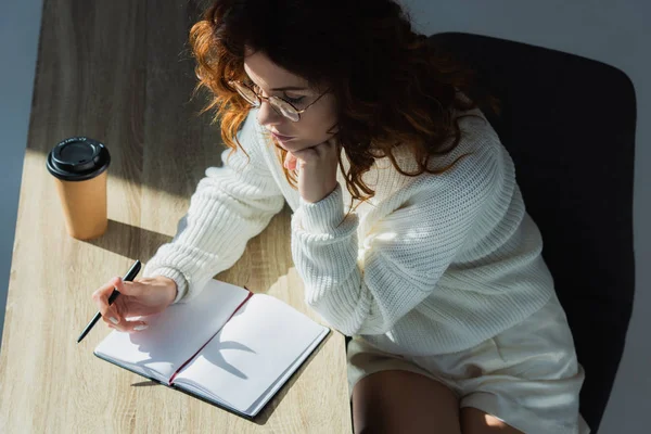 Overhead-Ansicht der nachdenklichen rothaarige Frau in Brille hält Stift in der Nähe leeres Notizbuch auf grau — Stockfoto