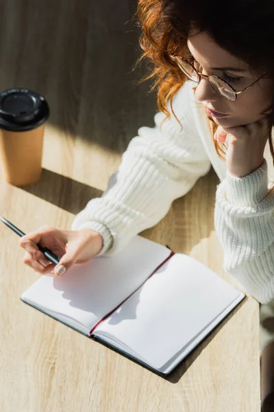 Overhead-Ansicht einer nachdenklichen rothaarigen Frau mit Brille, die einen Stift in der Nähe eines leeren Notizbuchs hält — Stockfoto