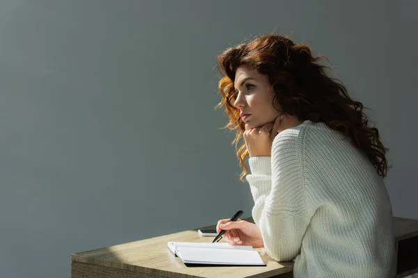 Dreamy curly redhead girl holding pen near notebook on grey — Stock Photo