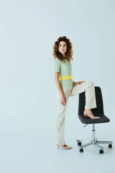 Attrayant rousse jeune femme debout près de chaise de bureau noire sur gris — Photo de stock