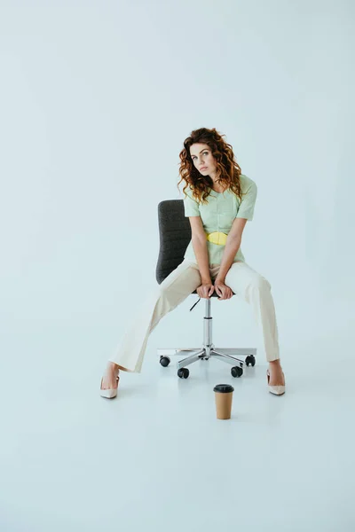 Attractive redhead young woman sitting on black office chair near paper cup on grey — Stock Photo