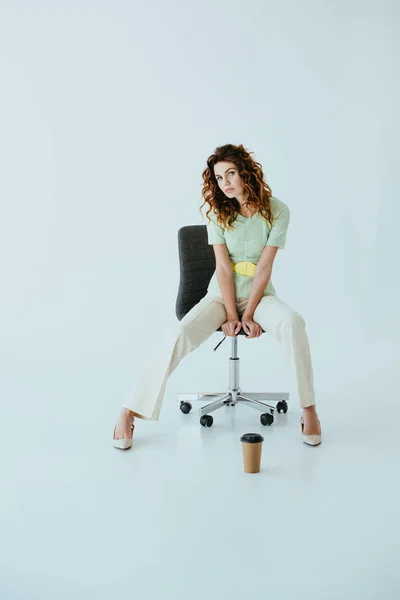 Attrayant rousse jeune femme assise sur chaise de bureau noire près de tasse en papier sur gris — Photo de stock