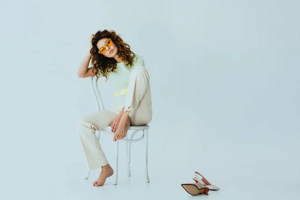 Curly redhead young woman in yellow sunglasses looking at camera while sitting on chair near heels on grey — Stock Photo