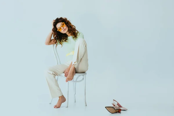 Rousse bouclée jeune femme en lunettes de soleil jaunes regardant la caméra tout en étant assis sur la chaise près des talons sur gris — Photo de stock