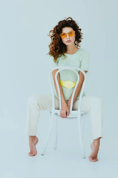 Curly redhead young woman in yellow sunglasses sitting on chair and looking at camera on grey — Stock Photo