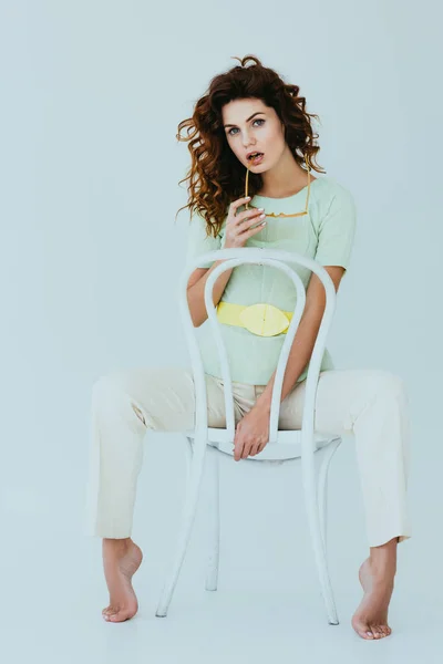Rousse bouclée jeune femme tenant des lunettes de soleil jaunes et assis sur la chaise sur gris — Photo de stock
