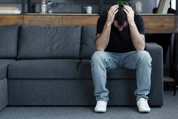 Sad adult man in blue jeans sitting on grey sofa and holding hands on head — Stock Photo