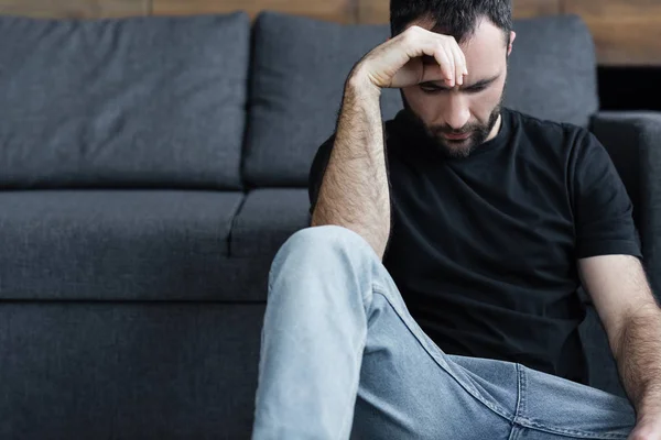 Depressed handsome man in black t-shirt sitting on floor and holding hand near head — Stock Photo