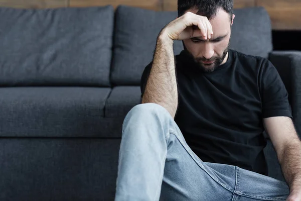 Hombre guapo deprimido en camiseta negra sentado en el suelo y sosteniendo la mano cerca de la cabeza - foto de stock