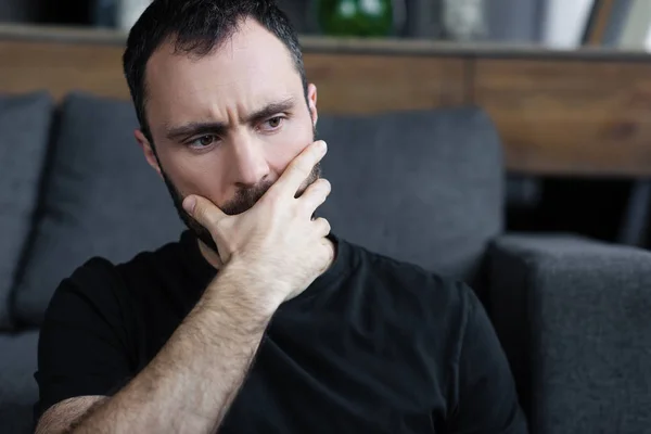 Homem sério em t-shirt preta segurando a mão na boca enquanto sentado em casa — Fotografia de Stock