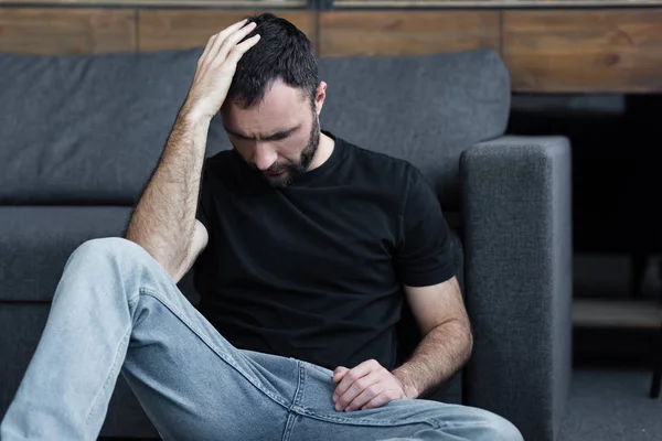 Homme barbu déprimé assis sur le sol près du canapé gris et tenant la main sur la tête — Photo de stock