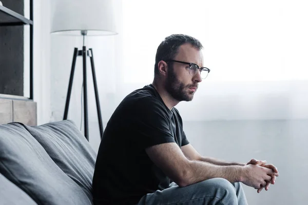 Nachdenklicher gutaussehender Mann mit Brille sitzt auf grauem Sofa und schaut weg — Stockfoto