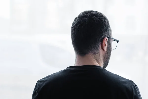 Vista trasera del hombre depresivo adulto en camiseta negra mirando por la ventana en casa - foto de stock