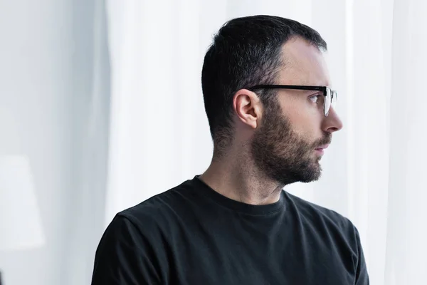 Hombre pensativo guapo en gafas mirando por la ventana en casa - foto de stock