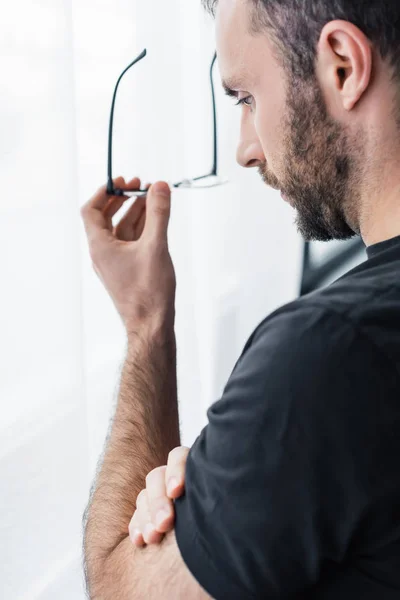 Sad bearded man holding glasses while standing by window and looking down — Stock Photo