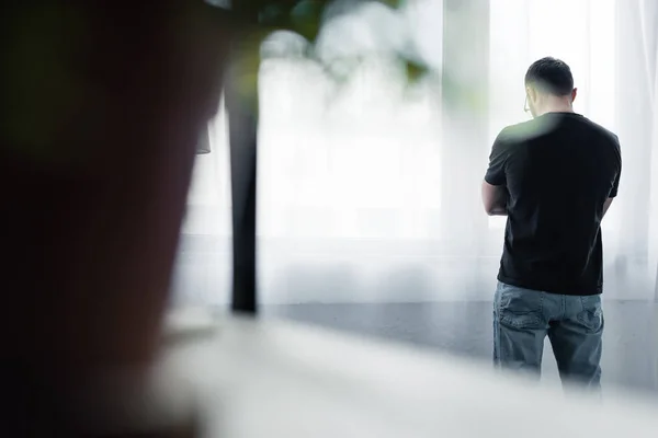 Selective focus of depressed man in black t-shirt standing by window at home — Stock Photo
