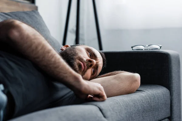 Depressed man lying on grey sofa at home and looking away — Stock Photo