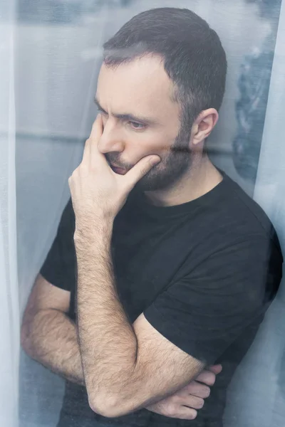 Hombre guapo y pensativo en camiseta negra de pie junto a la ventana y cogido de la mano en la boca - foto de stock