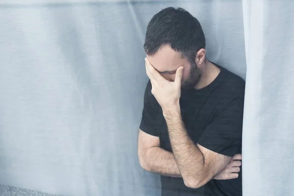 Homem solitário em t-shirt preta em pé pela janela em casa e segurando mão no rosto — Fotografia de Stock