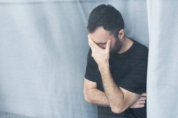 Homme seul en t-shirt noir debout près de la fenêtre à la maison et tenant la main sur le visage — Photo de stock