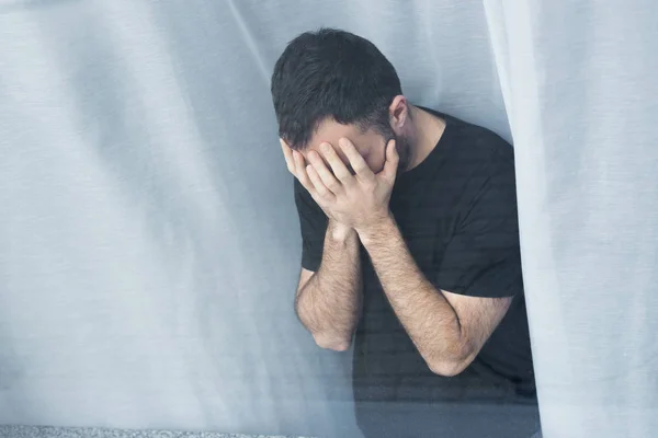High angle view of lonely man standing by window and holding hands on face — Stock Photo
