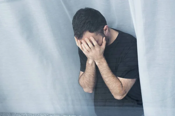 Vista de ángulo alto del hombre solitario de pie junto a la ventana y cogido de la mano en la cara - foto de stock